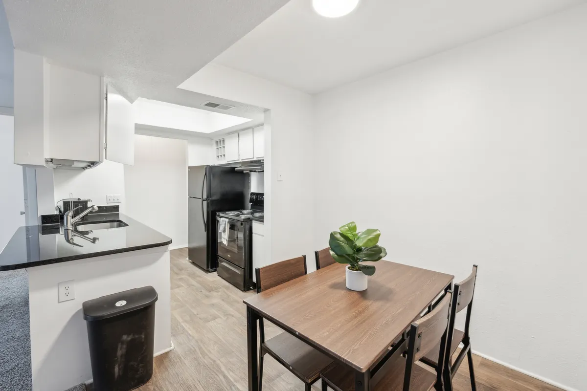 A close-up of the dining area showcasing a well-set dining table, adjacent kitchen counter with modern black appliances, and a vibrant potted plant. The scene glows with warm, natural light, suggesting a place where everyday meals and special moments are celebrated.
