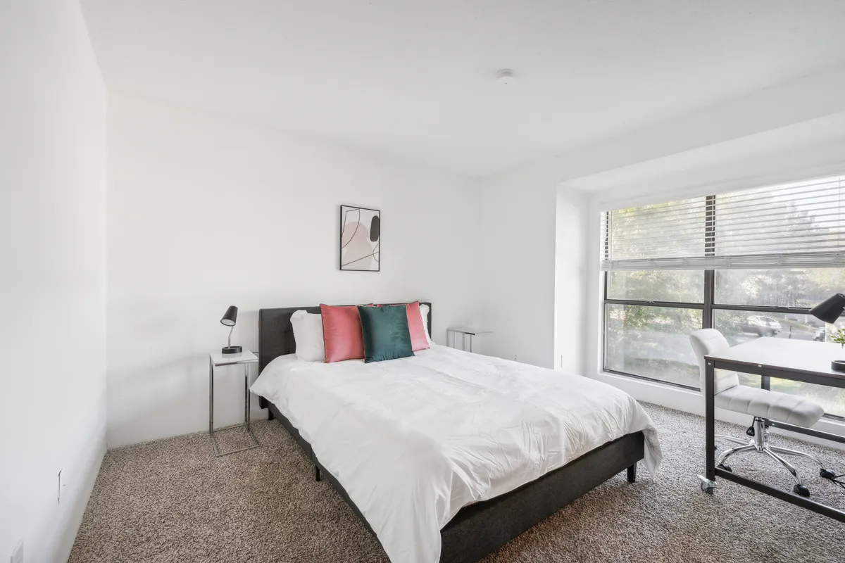 A bright bedroom with a bed adorned with colorful pillows, a desk and chair situated near a window, and abundant natural light filling the space. The setting offers both relaxation and a nook for productive work, making it ideal for guests who value both rest and functionality.