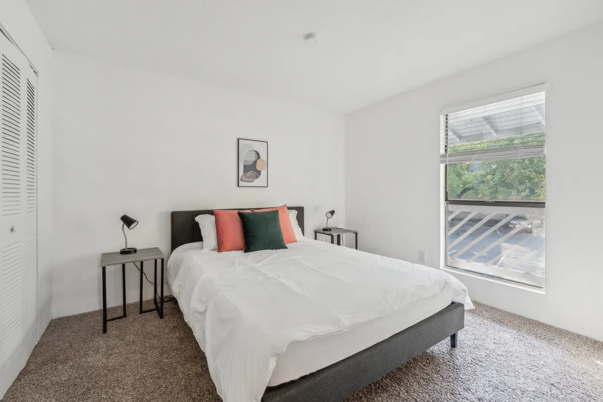 A tranquil bedroom featuring a neatly made bed with decorative pillows, stylish artwork on the wall, and nightstands with softly glowing lamps. Sunlight filters gently into the room, creating a cozy and inviting atmosphere ideal for rest and relaxation.